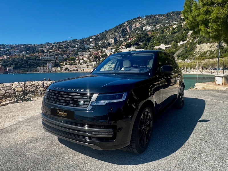 Range Rover Autobiography noir avec plaque Excellence au bord de la mer, face à une baie et des collines habitées.